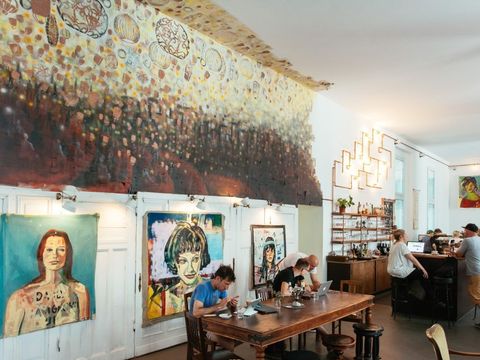 Im Hintergrund an der Wand mehrere Kunstwerke. Drei einzelne gemalte Bilder, die abstrakte Darstellungen von Frauenportraits zeigen. Davor sitzen an mehreren holzfarbenen Tischen und Stühlen Menschen, die Arbeiten. Vor einer Person steht ein Laptop. Rechts von den Tischen befindet sich der Bartresen. Auch hier stehen und sitzen mehrere Personen drumherum und unterhalten sich oder sind in die Arbeit vertieft.