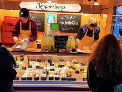 Käseverkaufsstand auf dem Wochenmarkt am Maybachufer. In der Auslage ganz viele verschiedne Sorten Käse, meistens im Stück. Dahinter stehen rechts und links zwei junge Männder, die den Käse verkaufen. Der Mann links im Bild trägt eine dunkelrote Fleecejacke und eine schwarze Mütze. Er hat eine gelbe Schürze um und nimmt gerade Geld aus einer Kasse. Der Mann rechts trägt einen dunkelblauen Pullover und ebenfalls eine gelbe Schürze. Er hat gerade ein Stück Käse aus der Auslage genommen. Im Vordergrund des Bildes sieht man drei Personen von hinten, die Käse kaufen wollen und gerade von den beiden Männern bedient werden. Auf der linken Bildseite eine einzelne Frau mit blonden Haaren, die geflochten sind. Sie trägt eine dunkle Winterjacke und einen Schal. Auf der rechten Bildseite eine Frau und ein Mann. Die Frau hat lange, braune, wellige Haare, die sie offen trägt. Sie hat eine dunkle Jacke an und über der rechten Schulter einen grünen Ricksack.Rechts neben ihr ein Mann mit einer Brille und braunen kurzen Haaren.Er trägt eine graue Jacke