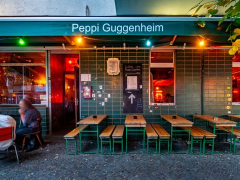 Auf der rechten Seite des Bildes sind mehrere Bierbankgarnituren aufgestellt. Auf der linken Seite sitzt ein Mann auf einem Stuhl an einem Tisch. Die Tische und Stühle stehen vor dem Ladenlokal "Peppi Guggenheim" Eine grüne Markise ist ausgefahren über die Tische und Stühle. In weißer Schrift steht hier der Name der Lokalität noch einmal drauf. Es hängen bunte Lampen unterhalb der Markise.