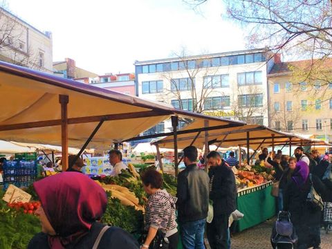 Mehrer Marktstände in einer Reihe. Im Bildvordergrund eine Frau mit einem dunkelroten Kopftuch