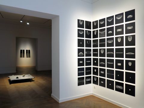 Man sieht einen Vorraum. Auf der rechten Seite eine Wand. Darauf eine Kunstinstallation, die aus vielen einzelnen kleinen schwarzen Bildern besteht, die zusammen ein großes Quadrat bilden. Auf den schwarzen Bildern sind helle abstrakte Formen zu sehen. Im nächsten Raum eine weitere Kunstinstallation auf dem Boden. Ein flaches graues Quadrat, das leicht erhoben steht. Darauf zwei Steine, die sich gegenüber liegen.