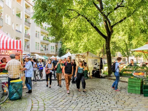 Eine kopfsteingepflasterte Straße mit Marktständen rechts und links. In der Mitte sind viele Personen zu sehen, die auf der Straße entlang laufen oder an einem der Marktstände stehen
