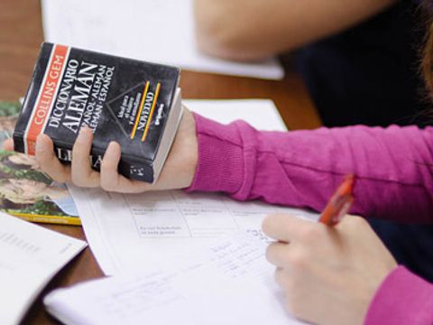 Eine Person hält in der einen Hand einen Stift und in der anderen ein Buch mit der Aufschrift ‚Diccionario Alemán‘