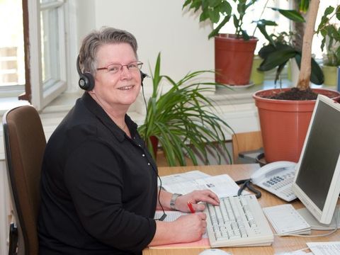 Frau sitzt am Computer mit einem Headset und schaut in die Kamera.