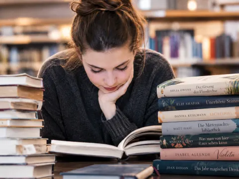 Junge Frau sitzt am Tisch in einer Bibliothek zwischen Bücherstapeln und liest