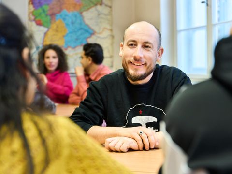 Teilnehmender eines Deutschkurses am Tisch sitzend im Unterrichtsgespräch mit anderen Teilnehmenden.
