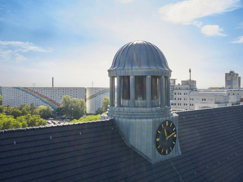 Blick auf die Turmuhr und das Dach der VHS Lichteneberg. Im Hintergrund Wohnhäuser des Bezirks Lichtenberg deren Fasseden mit Regenbögen verziert sind.