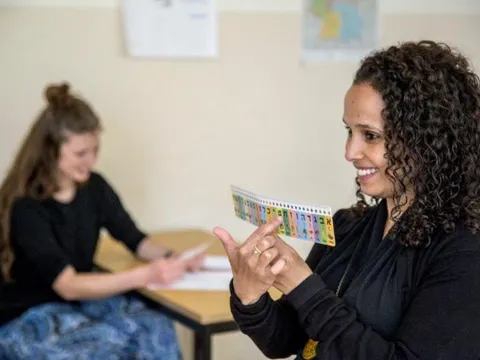 Zwei Frauen im Klassenzimmer; eine zeigt lachend eine farbige Karte, die andere sitzt am Tisch und schaut zu.