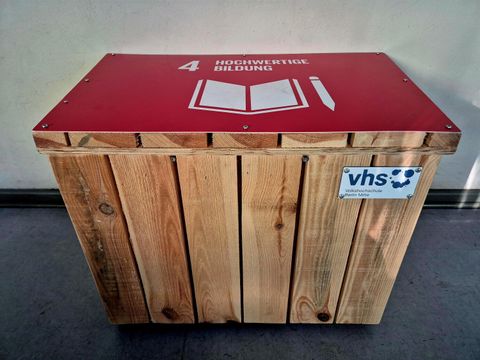 Sitzholzwürfel mit roter Oberseite und dem Symbol für ‚Hochwertige Bildung‘, beklebt mit einem VHS-Logo