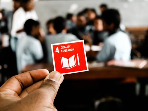 Eine Hand hält eine rote Karte mit weißem Mülltonnen-Symbol in die Kamera, im Hintergrund unscharf ein Klassenzimmer.