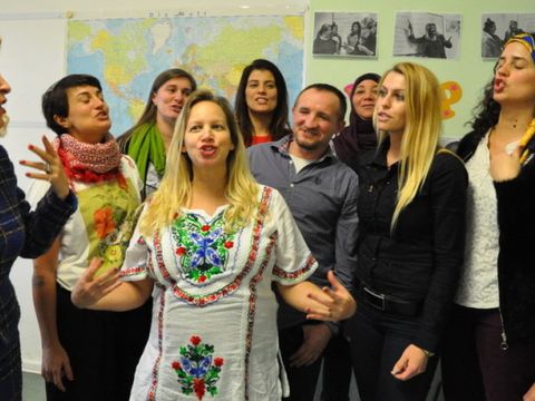 Eine Gruppe von Erwachsenen steht in einem Klassenzimmer und singt oder spricht gemeinsam. Im Vordergrund trägt eine Frau ein traditionell besticktes weißes Kleid. Im Hintergrund sind eine Weltkarte, Fotos und bunte Buchstaben an der Wand zu sehen.