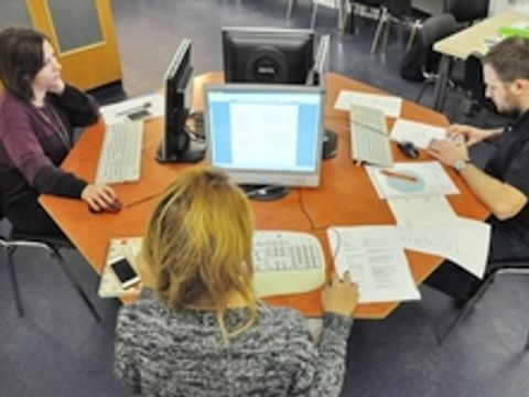 Personen sitzen an einem Tisch mit vielen Zetteln, alle arbeitet am Computer