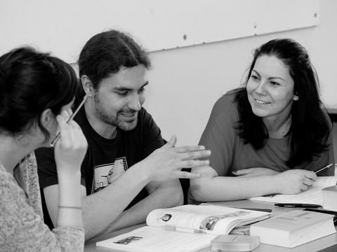 Drei Personen sitzen gemeinsam an einem Tisch, lächeln und unterhalten sich. Vor ihnen liegen Bücher und Lernmaterialien.