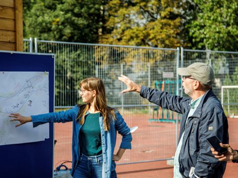 Zwei Personen stehen draußen auf einem Sportplatz, eine Frau zeigt auf eine Tafel