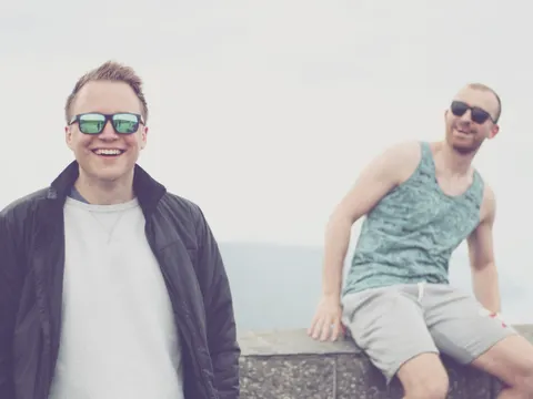 Zwei Männer mit Sonnebrille, einer sitzt auf Mauer