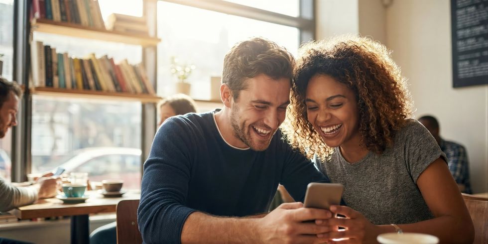 Zwei lächelnde Personen sitzen in einem Café und schauen gemeinsam auf ein Smartphone