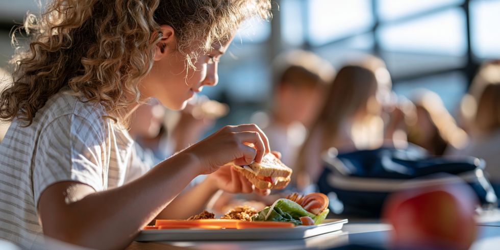Mädchen sitzt in hellem Raum am Tisch und isst ein Sandwich mit Gemüse auf Tablett