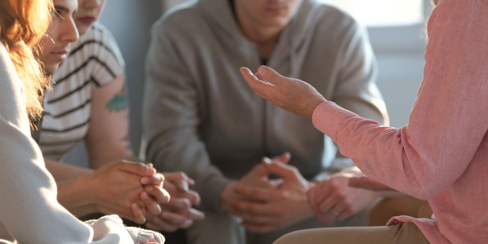 Eine Gruppe von Personen sitzt in einem Kreis und hält die Hände gefaltet. Eine Person spricht und gestikuliert mit einer Hand, während die anderen aufmerksam zuhören, typische Szene einer Gruppentherapie oder Besprechung.