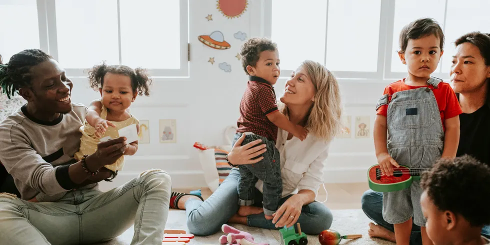 Mehrere Erwachsene und Kinder sitzen gemeinsam auf dem Boden eines hellen Raumes. Sie sind von buntem Holzspielzeug, einem Ball und Musikinstrumenten umgeben. Ein Kind sitzt auf dem Schoß einer Frau, während ein anderes Kind von einer weiteren Frau gehalten wird. Die Stimmung wirkt entspannt und freundlich. Im Hintergrund sind große Fenster und Wanddekorationen mit kindlichen Motiven zu sehen.