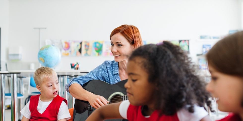 Schulkinder und Lehrerin mit Gitarre sitzen im Klassenzimmer, ein Kind mit geistiger Behindung