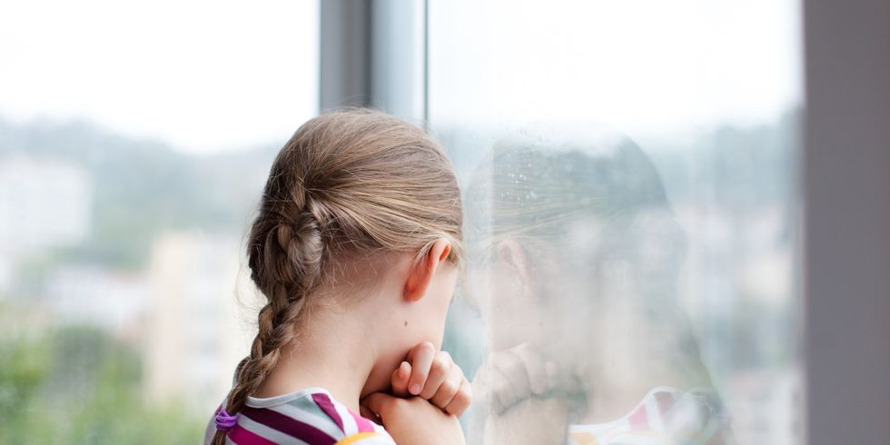 Sad child looking through rainy window at home. Upset kid in self isolation during quarantine.