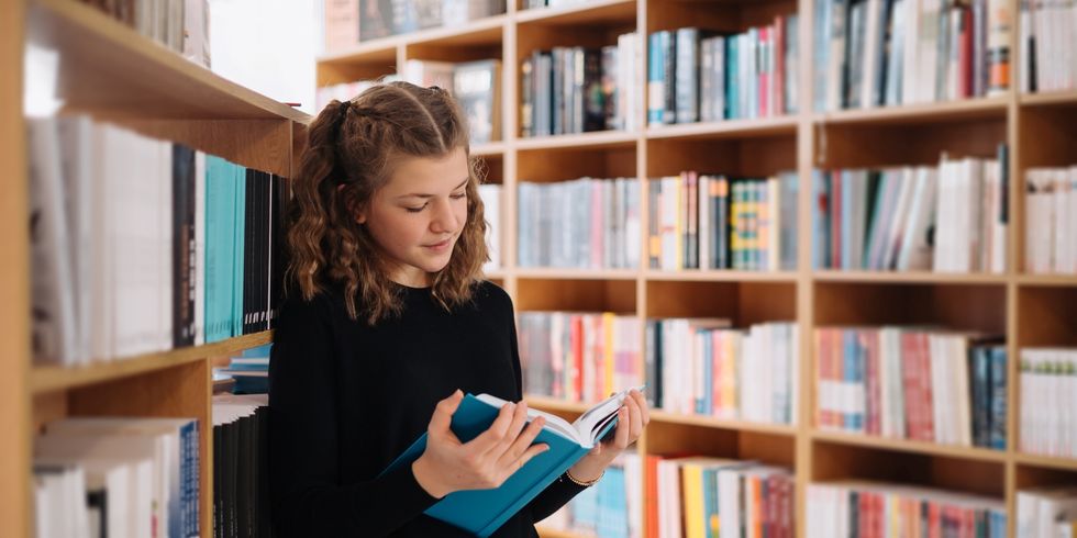 Jugendliche mit schwarzem Pullover liest in einem Buch in einer Bibliothek zwischen Bücherregalen