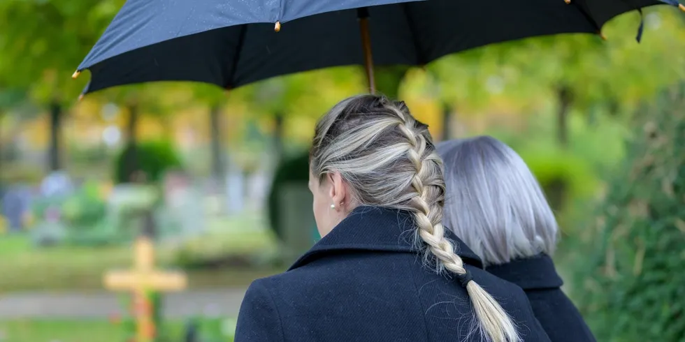 Zwei von hinten aufgenommene Personen mit dunklen Mänteln, eine Person hält einen Regenschirm, im Freien mit unscharfem Hintergrund aus Bäumen und Gräbern
