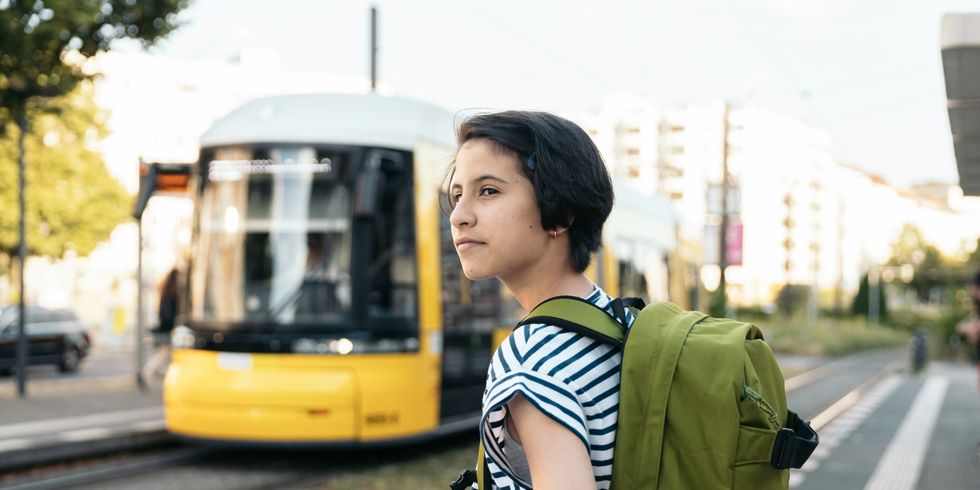 Person mit grünem Rucksack und gestreiftem T-Shirt steht an einer Straßenbahnhaltestelle vor einer gelben Straßenbahn in Berlin