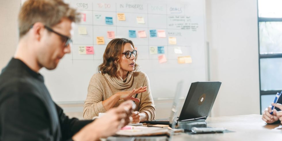 Frau mit Brille spricht an einem Tisch mit Laptop vor einer Wand mit bunten Notizzetteln, Mann im Vordergrund hat vor sich einen Laptop