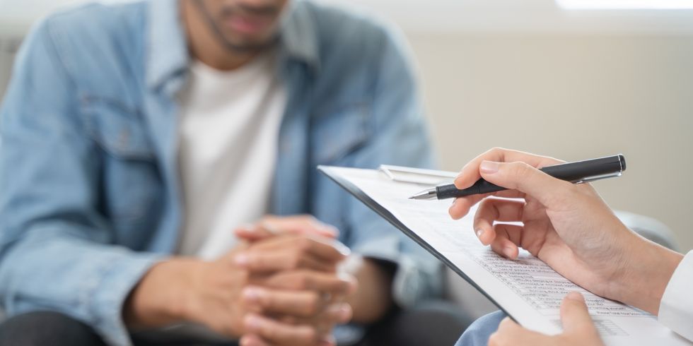 Person mit verschränkten Händen im Schoß sitzt gegenüber einer anderen Person, die einen Stift hält und auf einem Klemmbrett Notizen macht