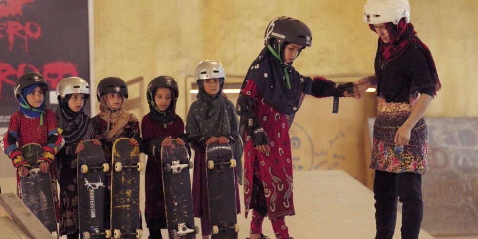 Mädchen steht auf einem Skateboard, weitere Mädchen halten im Hintergrund das Skkateboard in der Hand