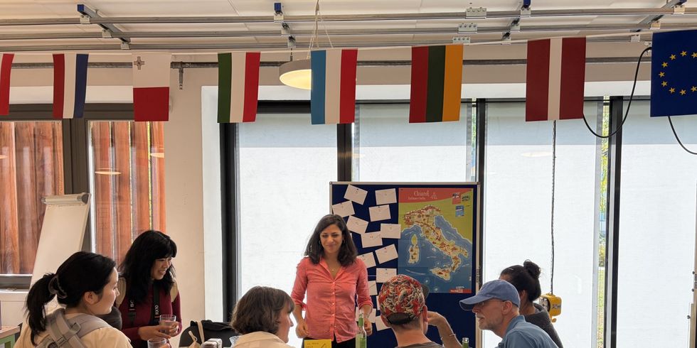 Bildbeschreibung: Eine Sprachlehrerin mit sechs Menschen an ihrem Tisch, die im Gespräch sind. Im Hintergrund eine Aufsteller mit Notizen und einer Italienkarte.
