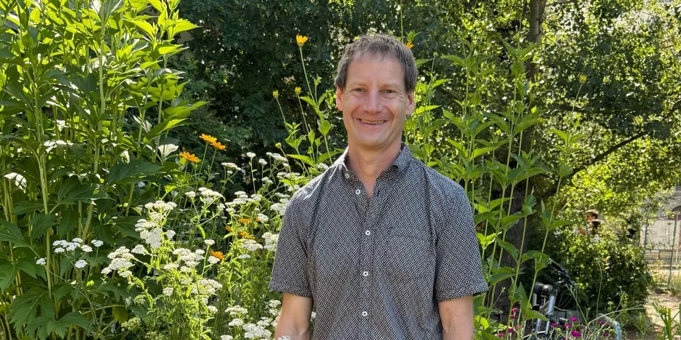 Ein Mann mit kurzem, braunem Haar steht vor hohen, blühenden Pflanzen in einem Garten. Er trägt ein kurzärmeliges, dunkelgraues Hemd mit feinem Muster und lächelt leicht. Im Hintergrund sind grüne Blätter, weiße und gelbe Blüten sowie ein Teil eines hellen Gebäudes zu sehen. Die Szene wirkt sommerlich und freundlich.