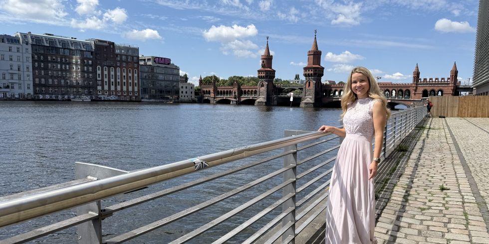 Eine Frau mit langen, blonden Haaren steht an einem Geländer am Ufer eines Flusses. Sie trägt ein helles, ärmelloses Kleid mit Spitzenoberteil und lächelt in die Kamera. Im Hintergrund ist die Oberbaumbrücke mit ihren markanten Türmen und einem gelben Zug zu sehen. Der Himmel ist blau mit wenigen Wolken.