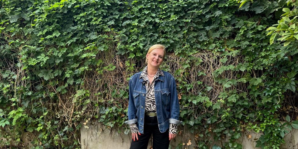 Eine Person steht vor einer dichten, grünen Efeuwand. Sie trägt eine blaue Jeansjacke, ein gemustertes Oberteil und eine schwarze Hose. Der Boden ist mit niedrigen Pflanzen bedeckt. Die Person blickt in die Kamera und lächelt leicht.