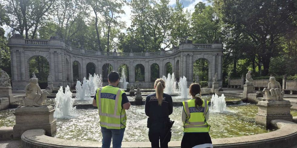 Drei Personen stehen mit dem Rücken zur Kamera vor einem großen, runden Brunnen mit mehreren Wasserfontänen. Sie tragen gelbe Warnwesten mit reflektierenden Streifen und dunkle Kleidung. Im Hintergrund ist eine steinerne Balustrade mit Bögen und Skulpturen zu sehen, dahinter hohe Bäume. Die Szene spielt sich bei Tageslicht ab, der Himmel ist teilweise bewölkt.