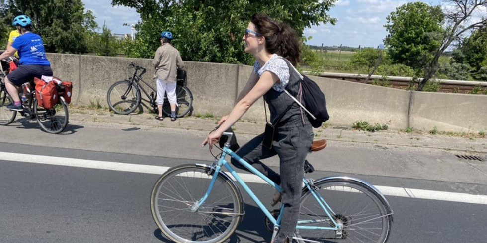 Eine Frau mit lockigen, dunklen Haaren fährt auf einem hellblauen Fahrrad auf einer asphaltierten Straße. Sie trägt einen schwarzen Rucksack, ein graues Oberteil und eine graue Hose. Im Hintergrund stehen zwei weitere Personen mit Fahrrädern an einer niedrigen Betonmauer, eine davon trägt einen blauen Helm und eine rote Fahrradtasche. Bäume und ein blauer Himmel mit wenigen Wolken sind sichtbar.