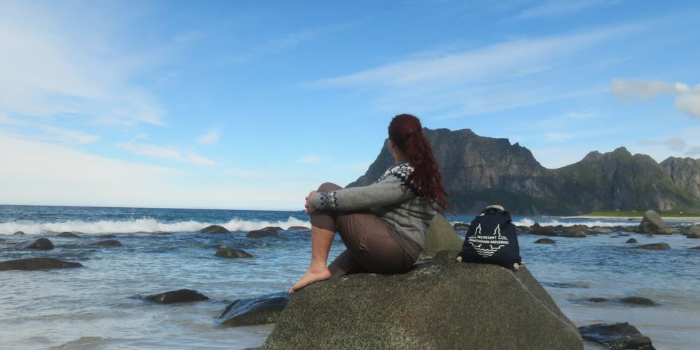 Eine Person mit langen, rötlichen Haaren sitzt barfuß auf einem großen Stein am Strand und blickt aufs Meer. Sie trägt eine graue Strickjacke mit Muster und braune Hose. Neben ihr steht ein dunkler Rucksack mit einem weißen Motiv. Im Hintergrund sind Wellen, ein heller Sandstreifen und eine markante, grüne Bergkette unter blauem Himmel zu sehen.