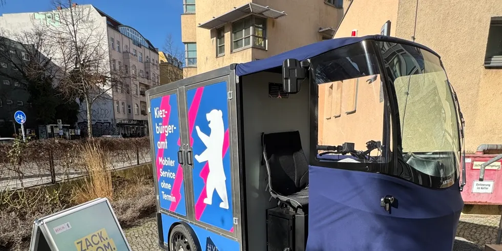 Lastenfahrrad mit Kofferaufbau mit blauem Werbeaufdruck und offenem Fahrersitz steht auf gepflastertem Gehweg neben einem aufgestellten Werbeschild