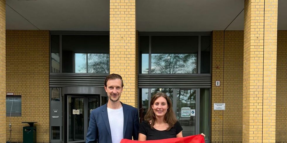 Bezirksbürgermeister Gordon Lemm und die QSF-Beauftragte Vanessa Krah stehen mit einer Regenbogenfahne vor dem Rathaus Marzahn-Hellersdorf. Im Hintergrund ist der Schriftzug "Rathaus" mit dem Bezirkswappen zu sehen