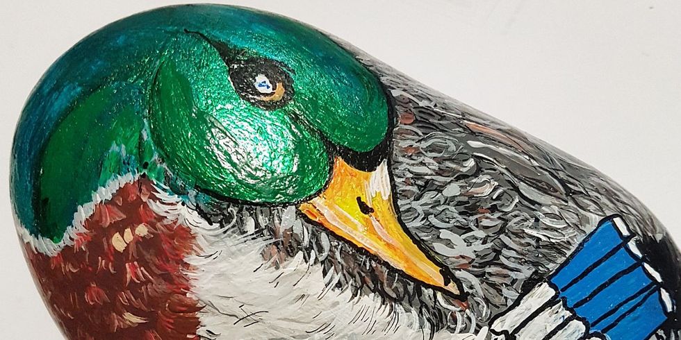 Das Bild zeigt einen Stein mit abgerundeter Form, bemalt mit dem Bild eines Erpels mit metallisch grünem Kopf, der seinen Hals auf den Körper geleget hat