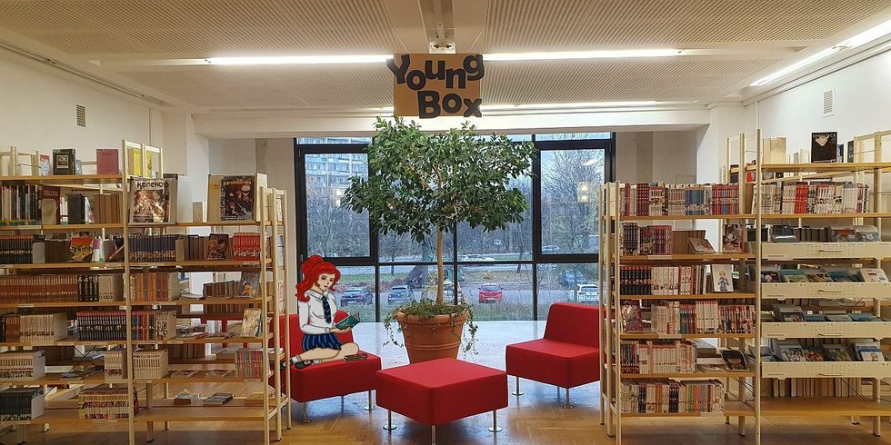 Jugendbereich der Mark-Twain-Bibliothek - Bücherregale, rote Hocker und Sessel und eine gezeichnete Mächenfigur, die mit untergeschlagenen Beinen auf einem der Sessel sitzt und in einem Buch liest