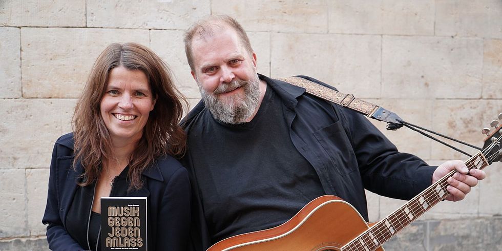 'Maria König' und 'Knut Schfleck' lachend vor einer Steinwand. Maria hält das Buch 'Musik gegen jeden Anlaß' in den Händen, Knut hat eine Akustikgitarre umghängt
