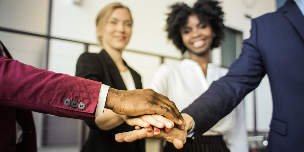 Eine Gruppe diverser Menschen stehen im Kreis und haben dabei ihre Hände übereinandergelegt