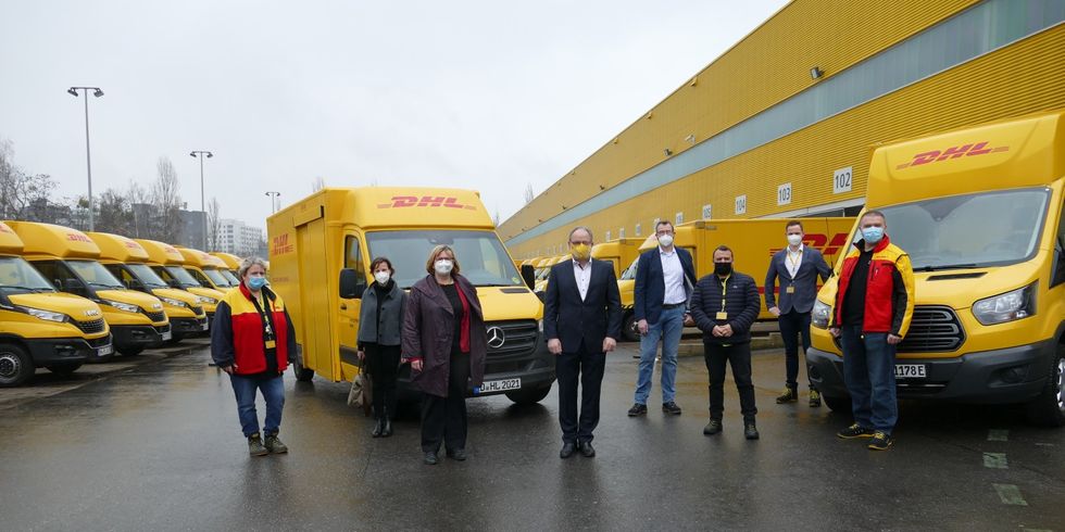 Menschen mit Maske stehen auf einem Gelände mit DHL Autos