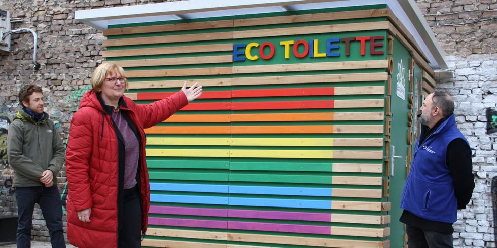 Drei Personen stehen vor einem mit regenbogenfarben bemalten Holzhäuschen mit der aufschrift Eco Toilette