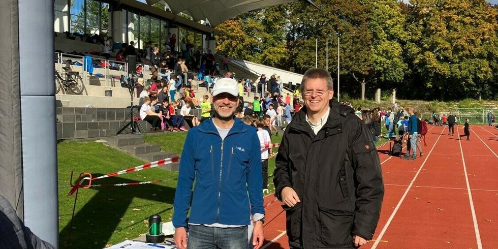 Zwei Männer stehen auf einer Laufbahn und lächeln in die Kamera. Im Hintergrund sitzen viele Menschen auf einer Tribüne.