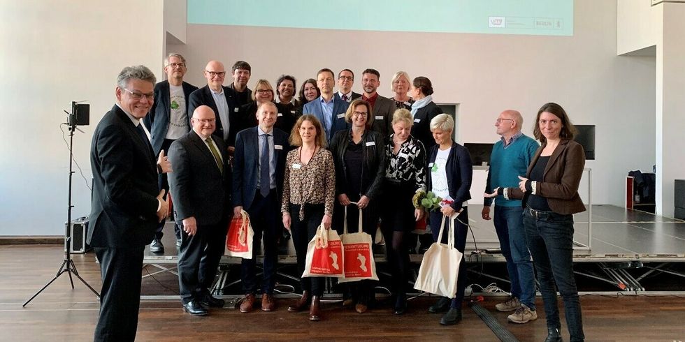 Eine Gruppe Menschen steht teils von und teils auf einer Bühne in einem großen Saal.