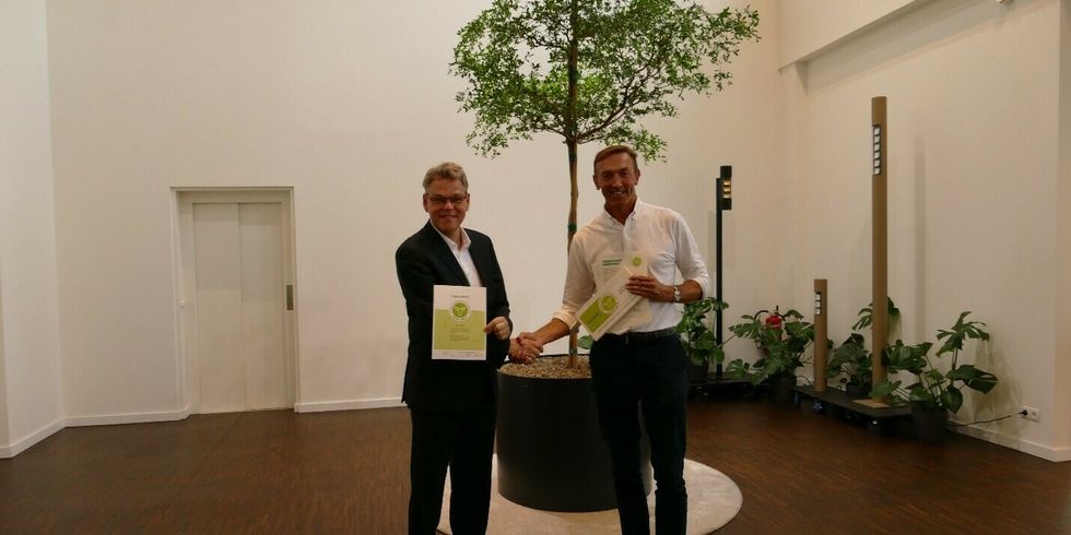 Zwei Männer stehen zusammen in einer Halle. Sie schütteln sich die Hand und halten Zettel mit dem Logo eines Grünen Hirschen hoch. Im Hintergrund sieht man einen kleinen Baum und andere Pflanzen mit Beleuchtung.