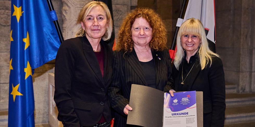 Drei Frauen stehen vor der Flagge der EU und der Berliner Flagge. Die mittlere Frau hält eine Urkunde in den Händen.